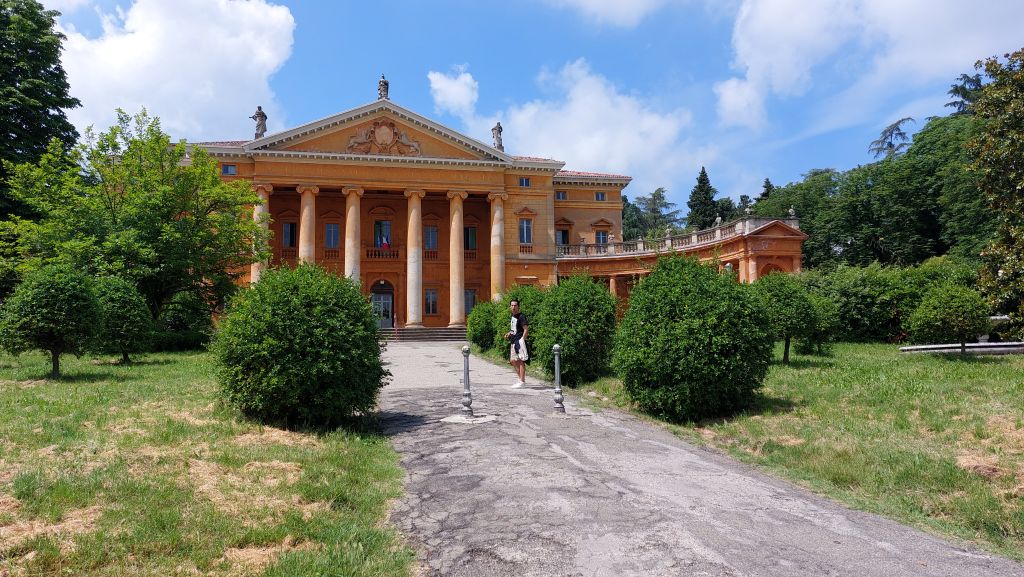 Una villa storica con colonne doriche e un grande giardino, con una persona che cammina lungo il sentiero.