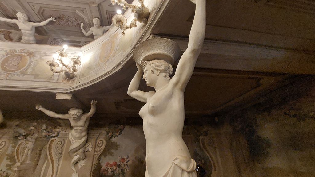 Statua di una figura femminile in pietra, con un vaso sopra la testa, all'interno di un ambiente decorato con stucchi e affreschi.