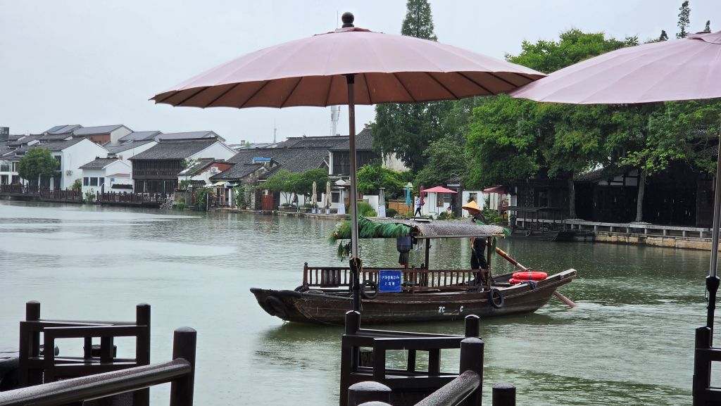 Una barca a remi naviga in un canale circondato da case tradizionali cinesi, mentre piove. Due ombrelloni rosa si trovano in primo piano.