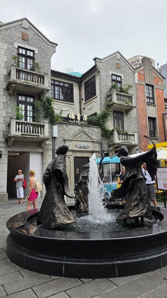 Fontana con sculture di bronzo che rappresentano figure danzanti, situata in una piazza urbana con edifici storici sullo sfondo.