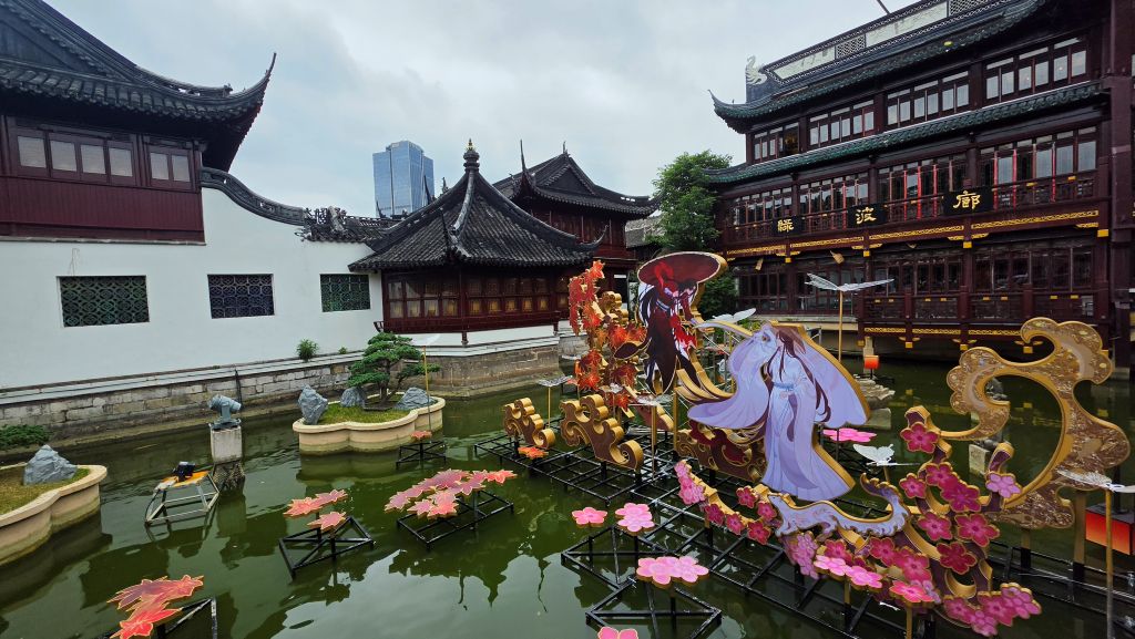 Vista di un giardino con acqua, caratterizzato da elementi decorativi colorati e personaggi stilizzati, sullo sfondo di edifici tradizionali cinesi.