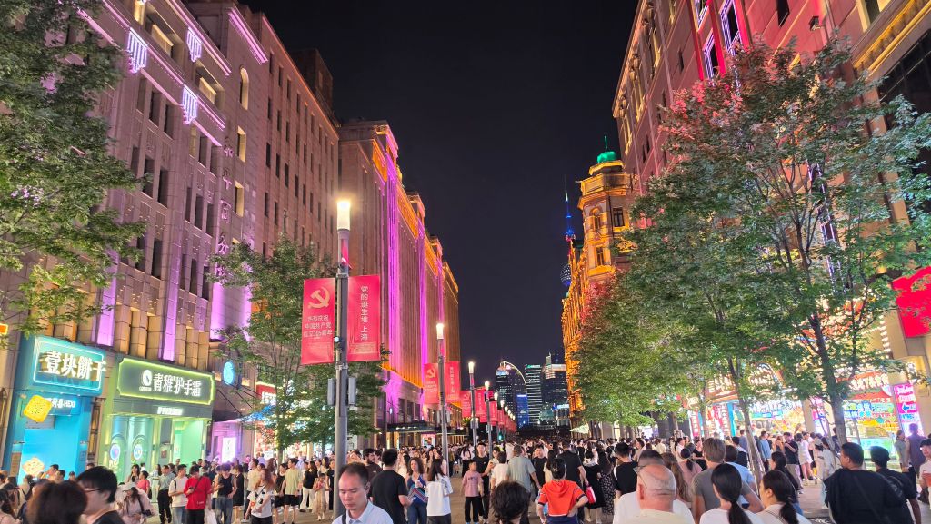 Una strada affollata di notte, costeggiata da alberi e negozi illuminati con luci colorate, a Shanghai.