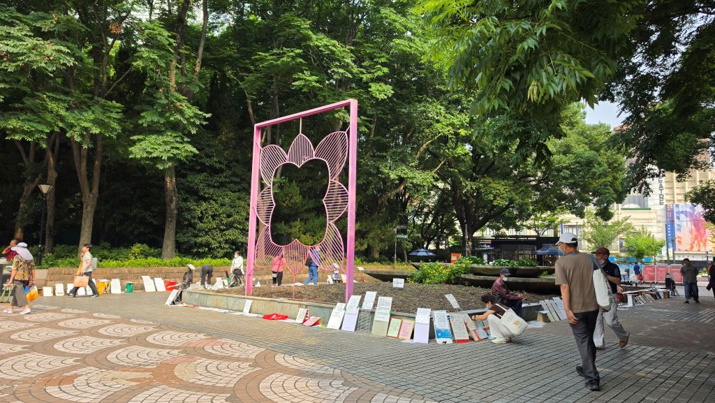 Un'area del parco con una grande struttura rosa a forma di fiore e visitatori che passeggiano. A terra ci sono vari cartelli esposti.