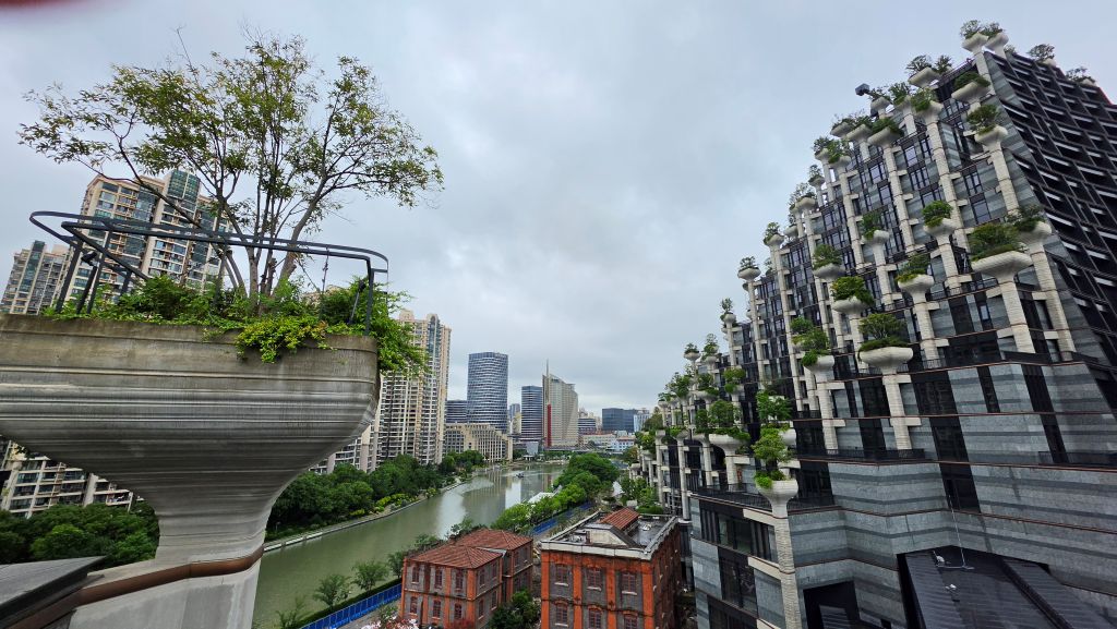 Vista panoramica di un paesaggio urbano con grattacieli moderni e vegetazione, comprese piante su balconi a strapiombo, lungo un fiume sotto un cielo nuvoloso.